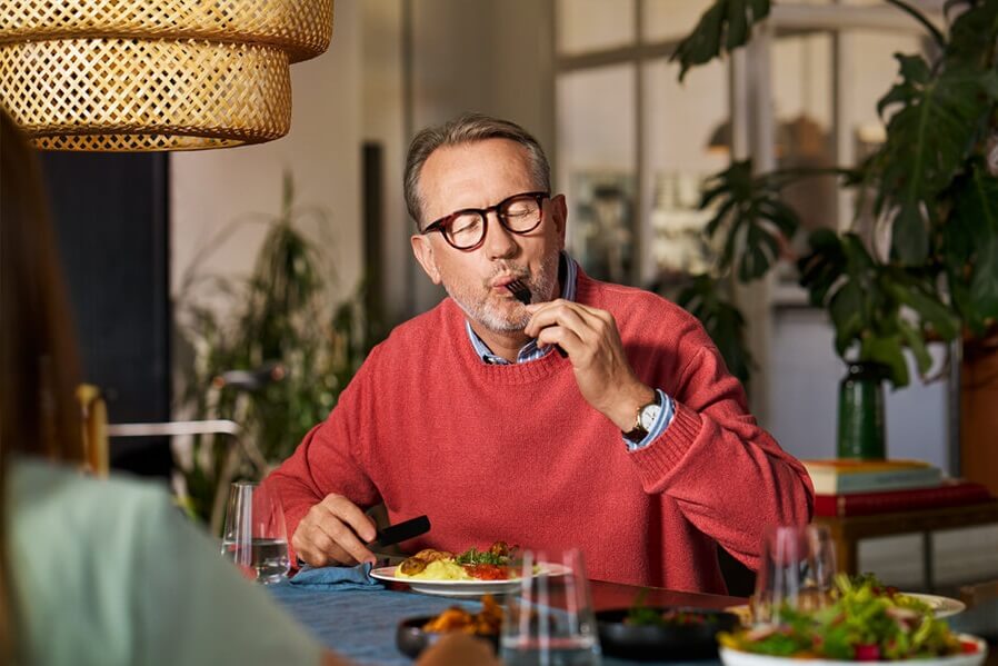Charmanter Mann beim Dinner beißt genussvoll von Gabel mit Hähnchen-Salat ab.
